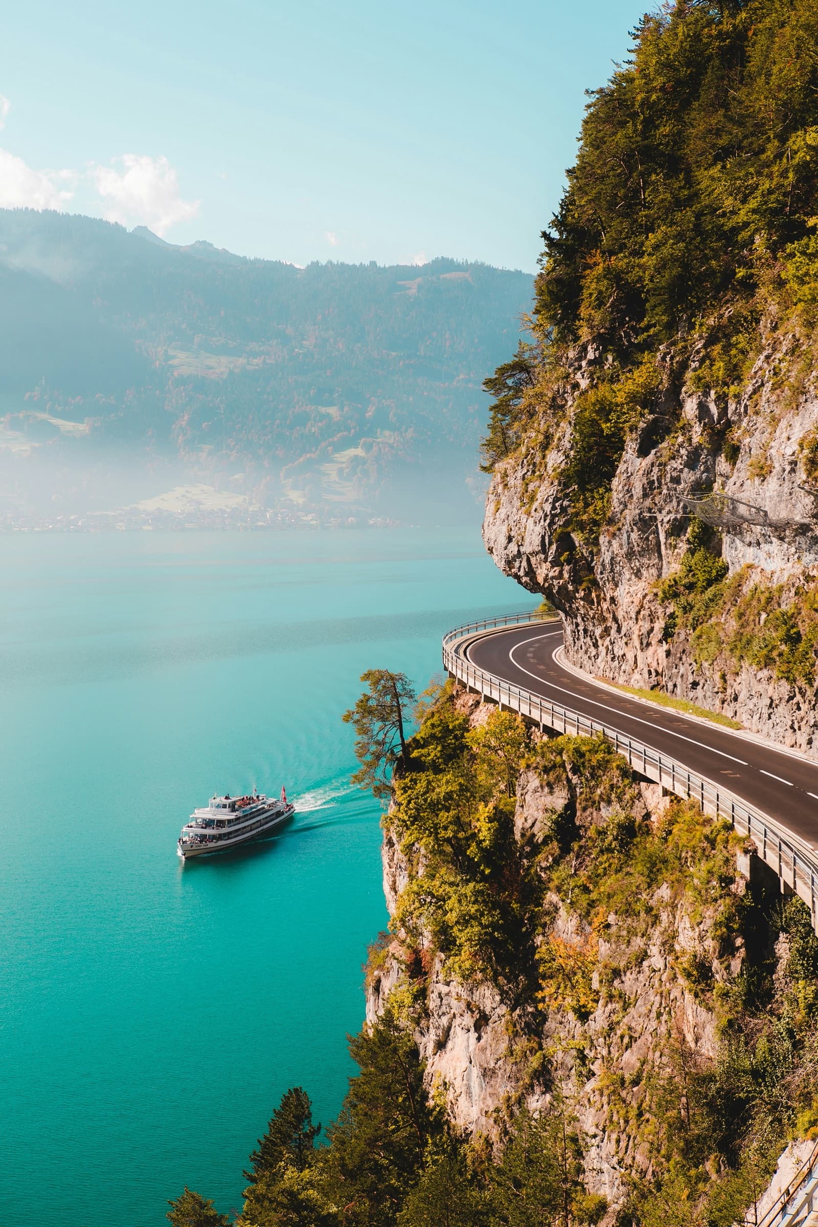 Mountain road curving along a cliffside above a turquoise alpine lake as a passenger ferry crosses the water below