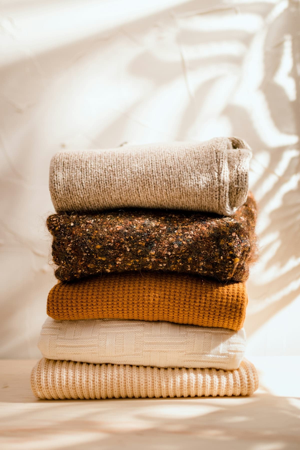 Stack of folded knit sweaters in oatmeal, mustard, and ivory under leaf shadows