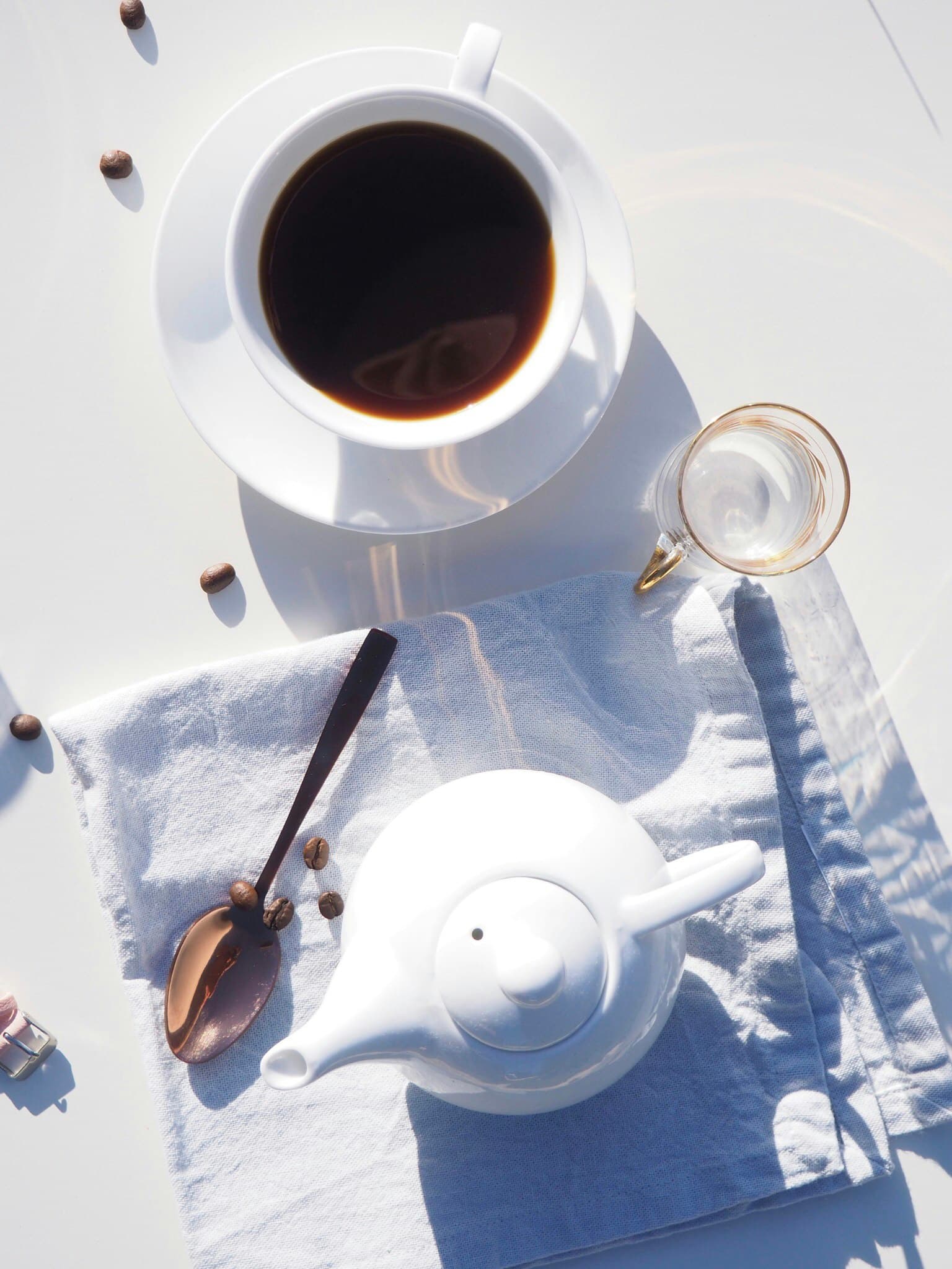 Morning coffee in a white cup beside a teapot on crumpled linen in bright sun